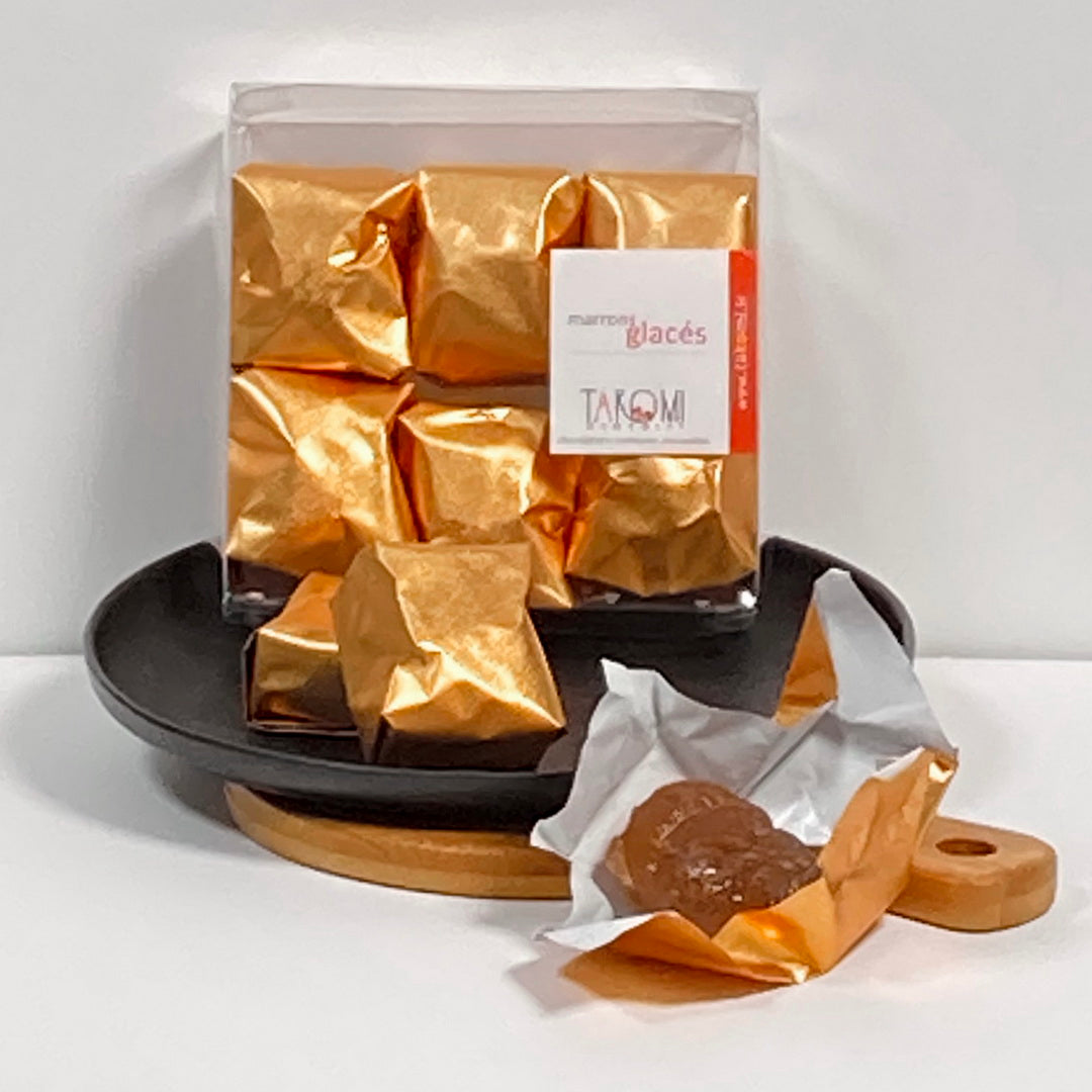 Marrons glacés emballés en papier doré dans un coffret noir, un marron confit déballé au premier plan